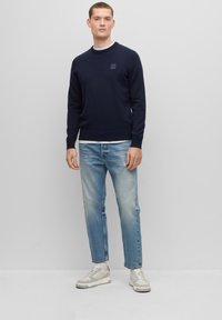 Donkerblauwe sweatshirt met ronde hals en klein logo-blik, gecombineerd met lichtblauwe versleten jeans en beige sneakers.