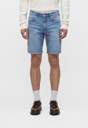 Mand iført lyseblå denimshorts, hvid langærmet skjorte, hvide sokker og brune lædersko, står mod en ensfarvet baggrund.