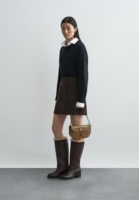 Pull noir, jupe en daim marron avec poches, bottes marron jusqu'aux genoux avec bord en fourrure, et sac à main beige avec clous. Design simple et moderne.