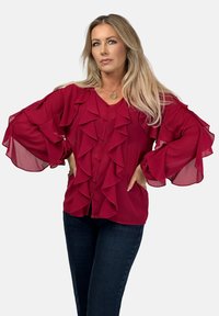 Rote Chiffonbluse mit Rüschendetails an der Vorderseite und den Ärmeln, mit einer Knopfverschluss-Design und einem lockeren Schnitt. Kombiniert mit dunklen Jeans.