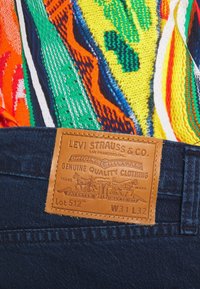 Denimjeans i en mörkblå tvätt har en baksida av brunt läder, präglad med varumärkes- och produktinformation, tillsammans med en färgglad mönstrad skjorta.