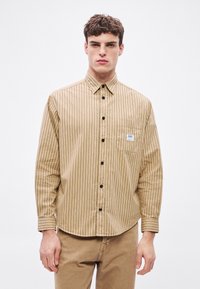 Dolga majica z dolgimi rokavi s tankimi navpičnimi črnimi črtami, z zadrgo na gumbke, ovratnikom in sprednjim žepom. Tkanina se zdi gladka s rahlo teksturo.