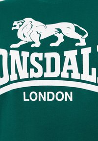 Zöld póló, amelyen egy fehér oroszlán grafika látható, a "LONSDALE" felirat merész betűkkel a "LONDON" szöveg felett. Pamut anyag, sima textúrával.