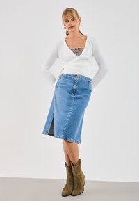 Weißer Wickelpullover, Jeansrock mit vorderem Schlitz und braunen Stiefeletten. Der Rock ist knielang mit einer blauen Waschung und fransigem Saum.