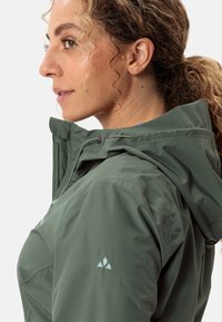 Giacca verde leggera con cappuccio, dotata di regolazioni con cordino e logo discreto sulla manica. Tessuto morbido e design aderente.