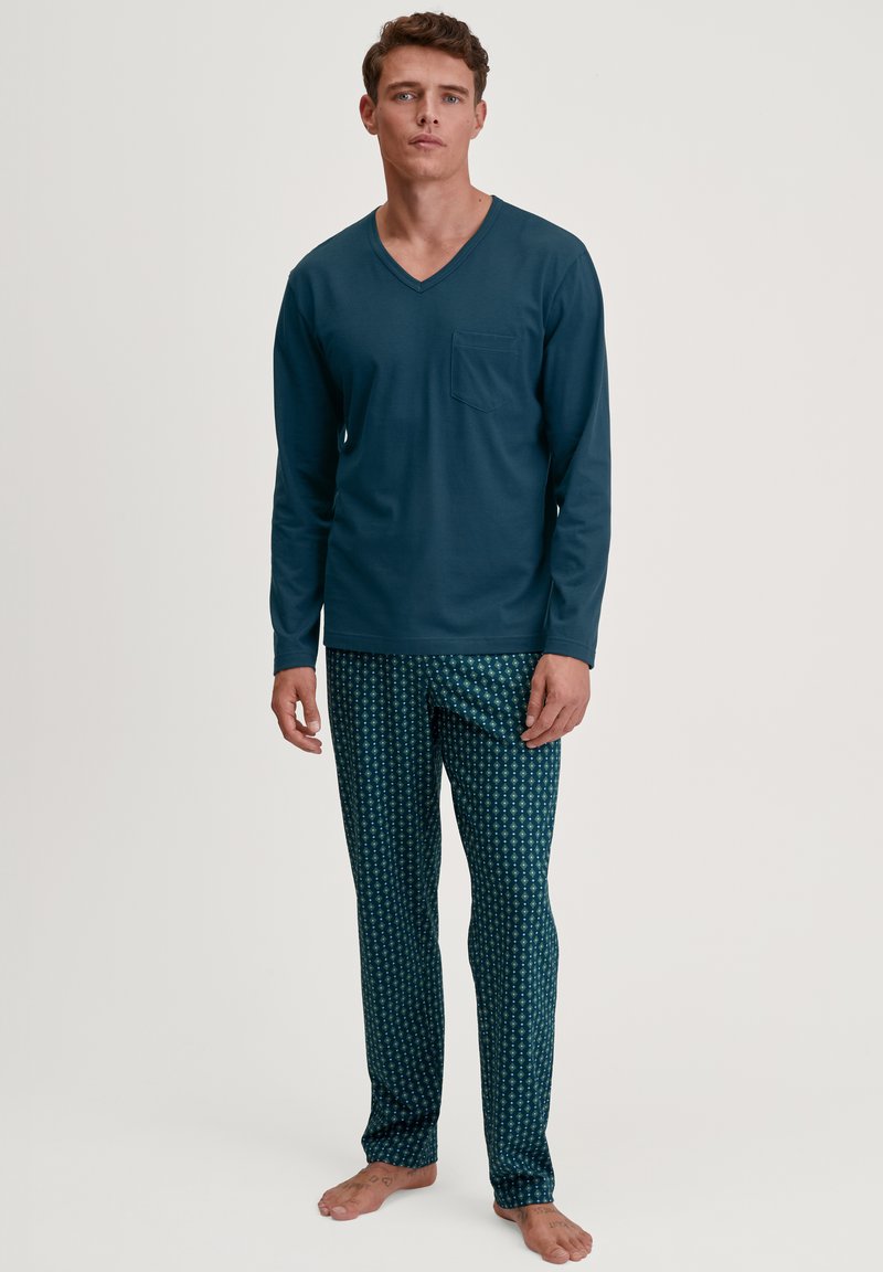 Haut à manches longues en V bleu marine avec poche, associé à un pantalon slim à motifs en vert foncé avec des designs floraux clairs.