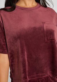 Bordeaux fluwelen t-shirt met een ronde hals, korte mouwen en een klein borstzakje. Zachte textuur met een lichte glans en een relaxte pasvorm.
