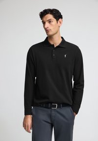 Svart långärmad polo med krage och tre knappar, med en liten vit logotyp på bröstet. Bärs tillsammans med grå byxor och ett bälte.