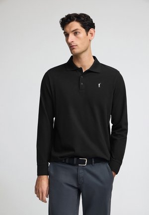 Polo a maniche lunghe nera con colletto e tre bottoni, con un piccolo logo bianco sul petto. Indossato con pantaloni grigi e una cintura.
