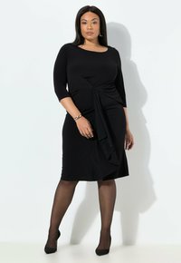 Vestido negro con mangas tres cuartos, que presenta un detalle en torsión en la cintura y un panel frontal fluido. Combinado con tacones altos negros y pantys.