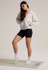 Sudadera de cremallera gris claro con cremallera dorada, pantalones cortos negros y calcetines tobilleros blancos con texto impreso, posados sobre una colchoneta de ejercicio beige.