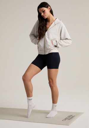 Sudadera de cremallera gris claro con cremallera dorada, pantalones cortos negros y calcetines tobilleros blancos con texto impreso, posados sobre una colchoneta de ejercicio beige.