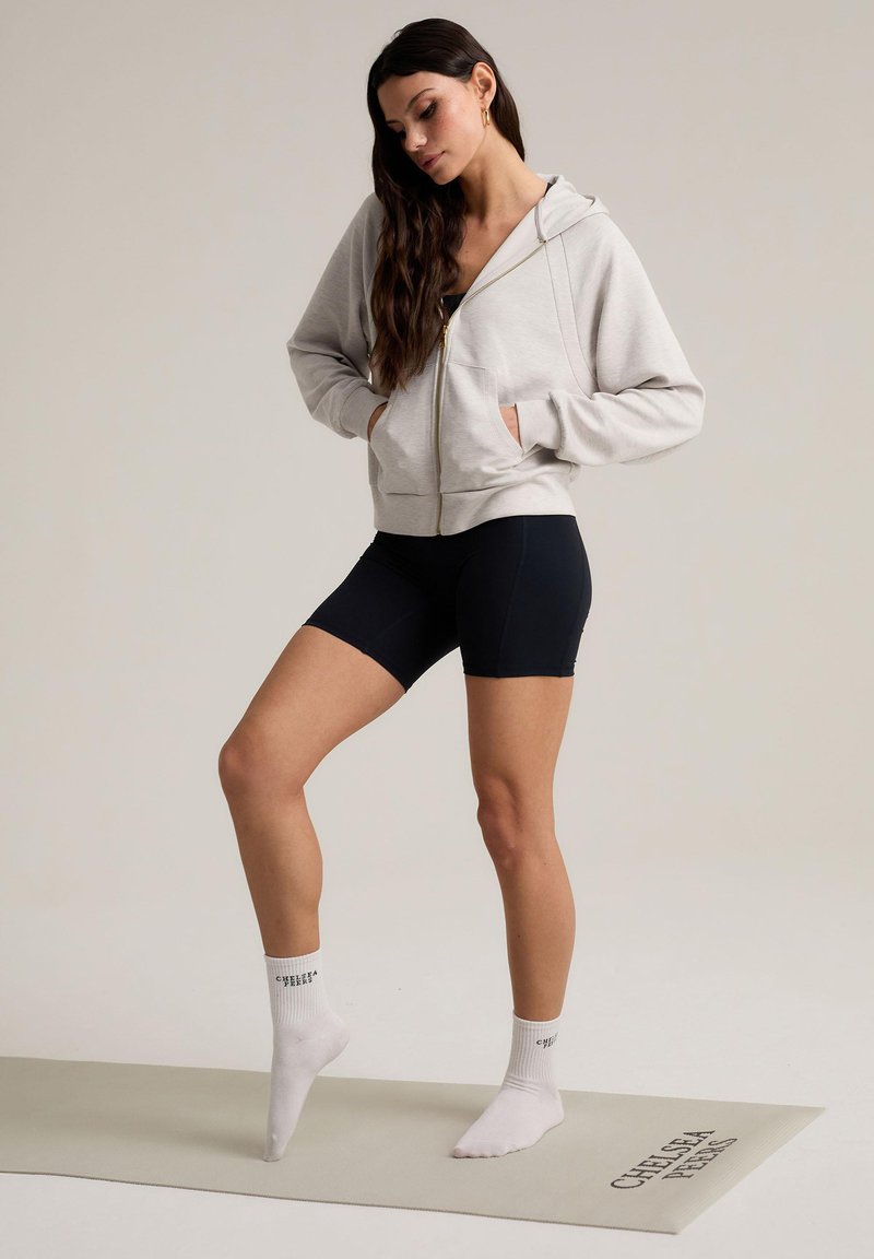 Sudadera de cremallera gris claro con cremallera dorada, pantalones cortos negros y calcetines tobilleros blancos con texto impreso, posados sobre una colchoneta de ejercicio beige.