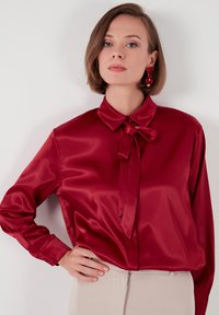 Red satin blouse with long sleeves, a collar, and bow detail at the neck. Smooth texture, fitted at the waist, paired with a light-colored bottom.