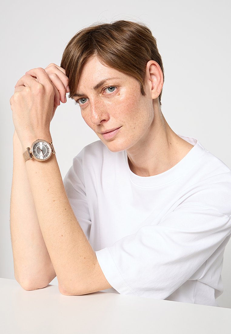 Kobieta z krótkimi brązowymi włosami, ubrana w białą koszulę, opiera ramiona na stole i prezentuje srebrno-złoty zegarek na nadgarstku.