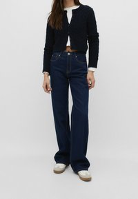 Mörkblå utsvängda jeans med ljus sömnad och hög midja, kombinerat med en svart stickad kort cardigan över en vit skjorta. Vita sneakers.