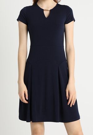 Vrouw draagt een marineblauwe jurk met korte mouwen, een keyhole-halslijn en een gouden stangdetail, staand tegen een effen lichte achtergrond.