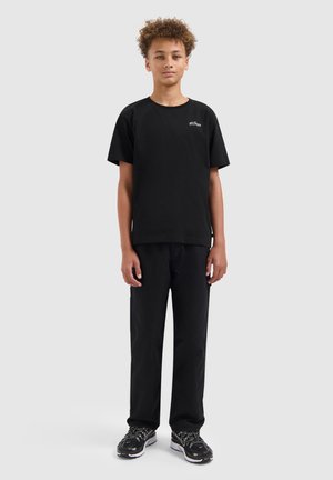 Tienerjongen met krullend haar, staand in een eenvoudig zwart T-shirt, zwarte broek en zwarte sneakers tegen een lichtgrijze achtergrond.