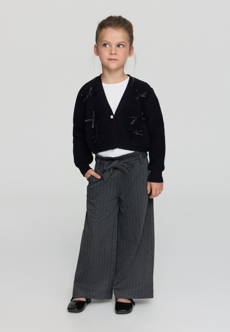 Cardigan corto nero con dettagli in nastro, abbinato a pantaloni larghi a righe grigie e scarpe basse nere. Camicia bianca sotto.