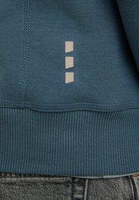 Ourlet en tissu côtelé vert sarcelle d'un vêtement avec une couture verticale et trois petites patches réfléchissantes rectangulaires beige alignées au centre.