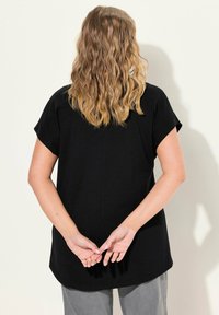Top negro de manga corta con un corte relajado y escote redondo. Presenta un detalle de costura en la parte posterior, combinado con pantalones grises claros. Ideal para ropa casual.