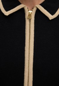 Vêtement en maille noire avec une bordure beige le long du col et de la fermeture éclair, avec un tirette doré gravé du nom de la marque Barbour.