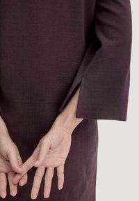 Hände hinter dem Rücken gefaltet, getragen wird ein dunkellila Langarmkleid mit einem seitlichen Schlitz am Handgelenk, vor einem schlichten hellen Hintergrund.