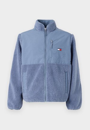Jachetă căptușită cu fleece de culoare albastru deschis, cu fermoar frontal, guler înalt, buzunare laterale și patch cu logo pe partea superioară stângă a pieptului.