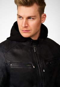 Zwarte leren jas met een rits aan de voorkant, twee ritszakken op de borst en een capuchon met trekkoorden. Soepele textuur en minimale hardware accenten.