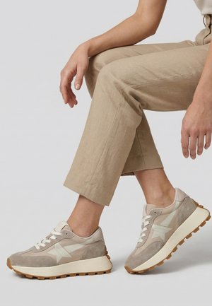 Osoba v béžových kalhotách a taupe teniskách s bílými hvězdnými akcenty, sedící se zkříženýma nohama na jednobarevném pozadí.