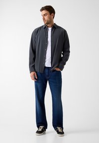 Donkergrijs overhemd over een wit t-shirt, gecombineerd met blauwe spijkerbroek en zwarte sneakers. De outfit heeft een casual, relaxte pasvorm.