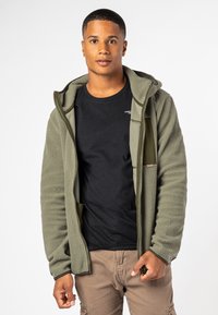 Fleece jack in olijfgroen met een ritssluiting aan de voorkant, zakken en een textuurstof. Draagt boven een zwart T-shirt, gecombineerd met beige broeken.