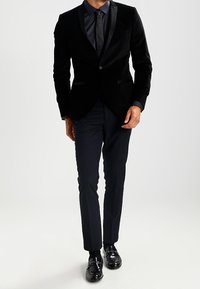 Black velvet tuxedo jacket, satin lapels, paired with dark trousers and shiny black shoes. Sleek design, tailored fit, minimal detailing.