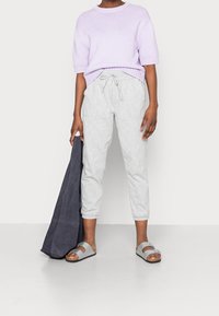 Osoba ubrana w jasnofioletowy sweter z krótkim rękawem, jasnoszare spodnie ze sznurkiem, szare sandały, trzymająca dużą ciemnoszarą torbę na ramię.