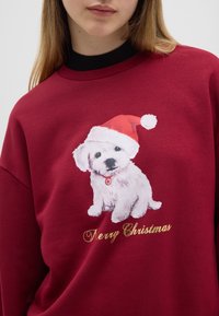 Bordeaux sweatshirt met een print van een witte puppy die een rode Santa-hat draagt, met daaronder "Vrolijk Kerstfeest" in goud. Zachte textuur.