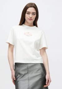 Biele bavlnené tričko s krátkymi rukávmi, okrúhlym výstrihom a stredovým grafickým logom v oranžovej farbe. Kombinované so sivou metalickou sukňou.