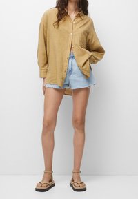 Chemise légère à manches longues de couleur moutarde avec des boutons, associée à un short en denim bleu clair effiloché et des sandales beiges.