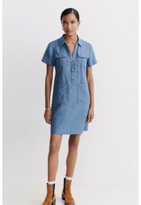 Abito camicia in chambray con maniche corte, chiusura con bottoni, due tasche sul petto, due tasche anteriori e orlo fino al ginocchio. Indossato con stivaletti alla caviglia.