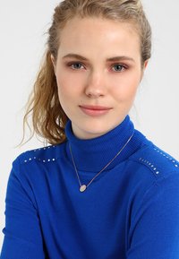 Maglione blu a collo alto con texture a costine, caratterizzato da borchie argentate sulle spalle, abbinato a una collana delicata e trucco naturale.