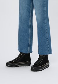 Czarne zamszowe wysokie botki z złotymi akcentami, z zamkiem błyskawicznym, gumową podeszwą i sznurowanym fasonem, połączone z niebieskimi dżinsami.