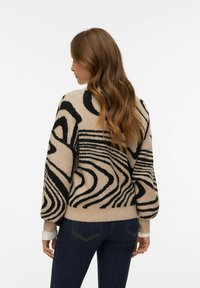 Béžový svetr s černými abstraktními vzory, s volnějším střihem a žebrovanými manžetami. Svetr má měkkou strukturu a výrazné designové prvky.