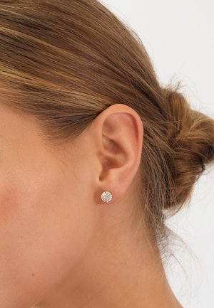 Round, clear crystal stud earring with gold-tone setting, showcasing a textured surface and four prong accents, worn in the ear.