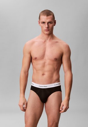 Ung mand med kort hår iført sorte Calvin Klein underbukser stående mod grå baggrund.