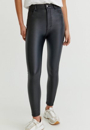 Femme portant des leggings noirs en similicuir taille haute et des baskets blanches, debout devant un fond uni.