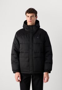 Homem vestindo uma jaqueta puffer preta com capuz e bolsos com fecho éclair, de pé contra um fundo claro liso.