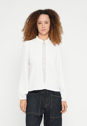 Λευκό μακρυμάνικο μπλουζάκι με βολάν στο γιακά, δαντέλα στο μπροστινό μέρος και πλισέ μανίκια. Φορεμένο με σκούρο, ψηλόμεσο τζιν παντελόνι.