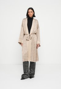 Kimono beige à motifs avec ceinture à nouer, doté de larges manches et d'un col contrastant. Associé à un pantalon noir à motifs et des chaussures pointues.