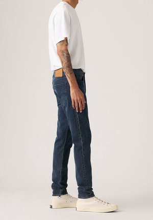 Homme portant un t-shirt blanc, un jean bleu foncé, des baskets montantes blanches et un tatouage visible sur le bras, debout de profil sur un fond uni.