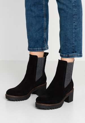 Sorte ruskind ankelstøvler med chunky hæle og mønstrede elastiske sidepaneler, båret med korte blå jeans.
