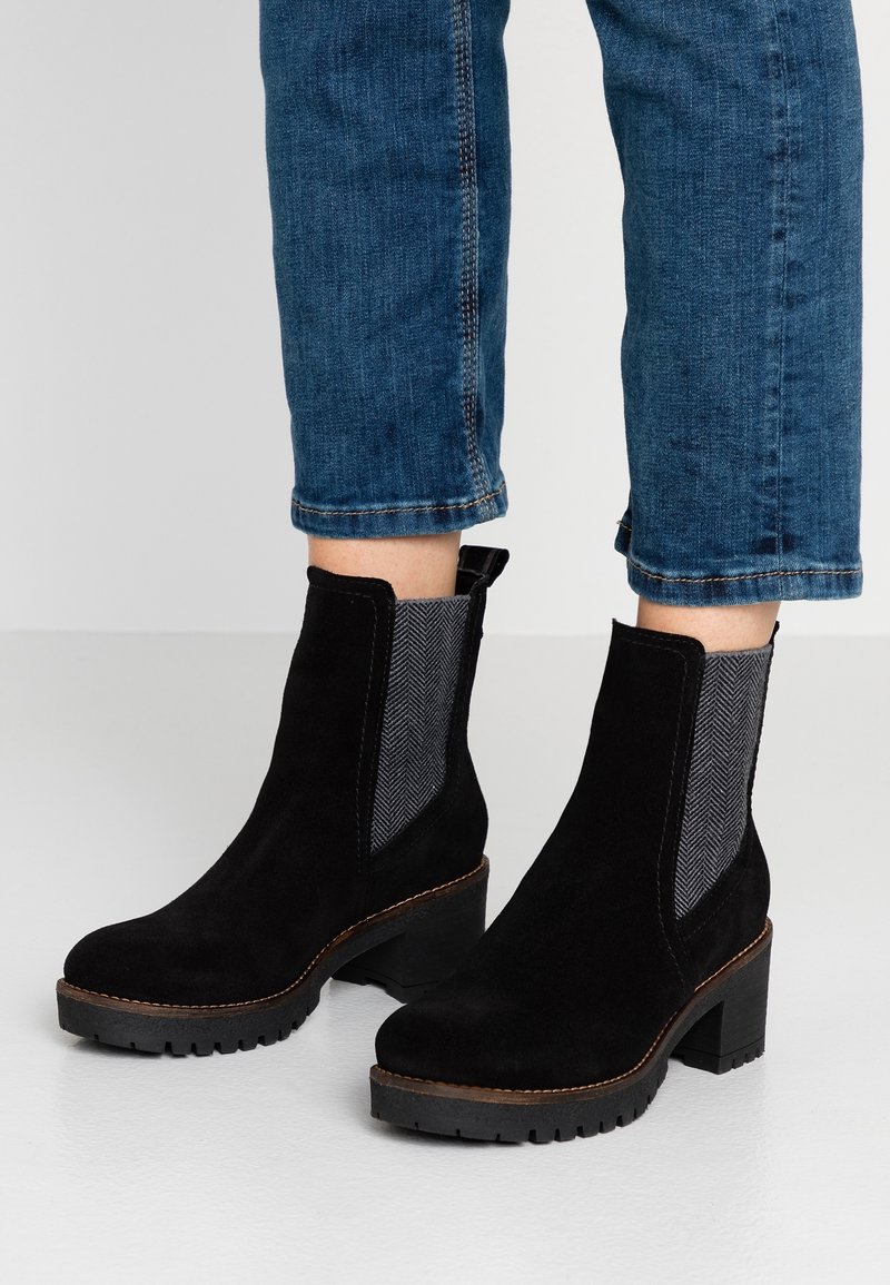 Bottines en daim noir avec talons épais et panneaux latéraux élastiques à motifs, portées avec un jean bleu raccourci.